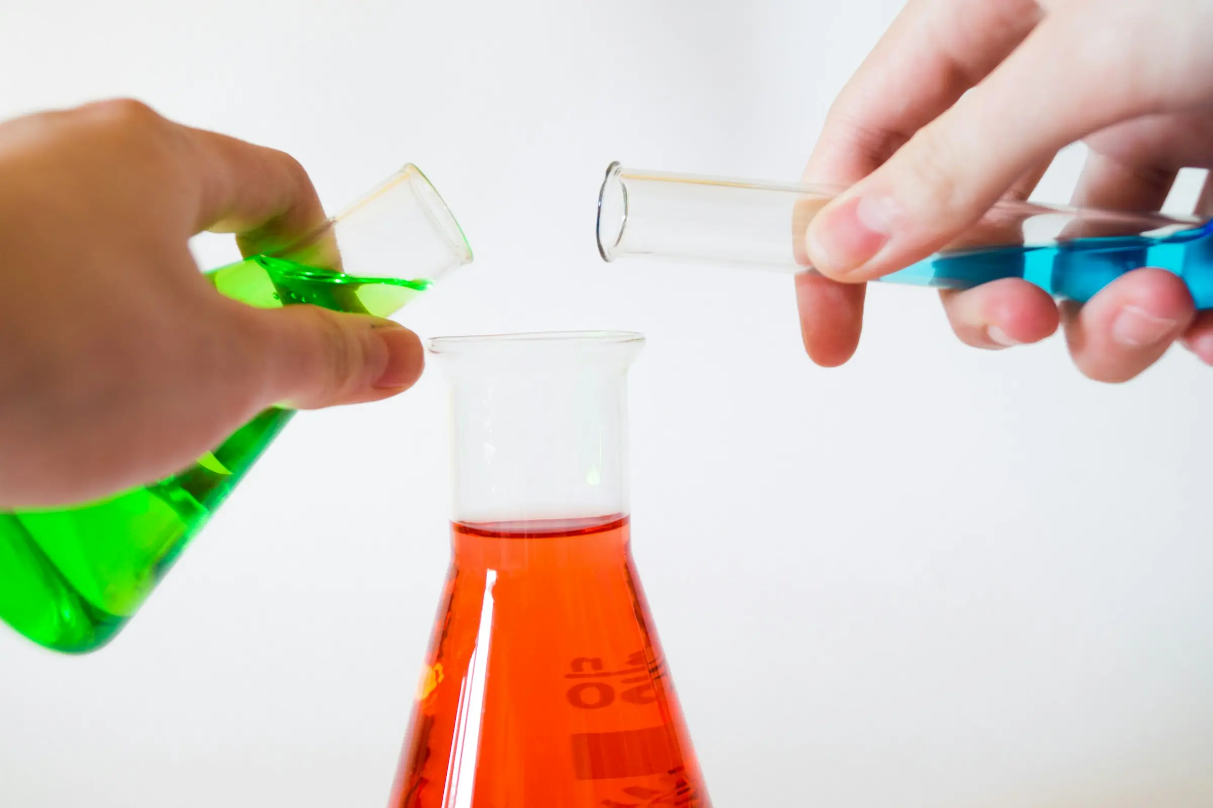 Hands combining colored solutions over laboratory glassware